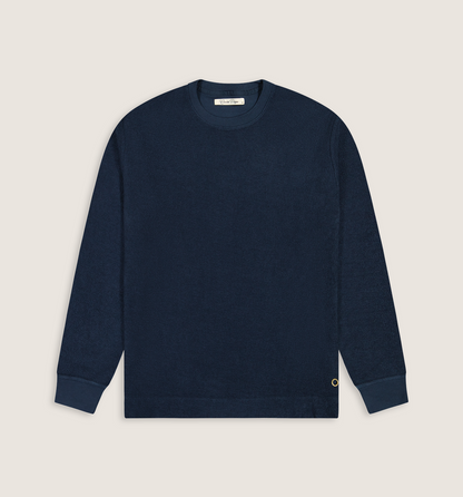 A dark navy blue crewneck sweatshirt with ribbed cuffs and hem, laid flat against a light beige background. A small gold embroidered 'C' logo is visible on the lower left cuff.