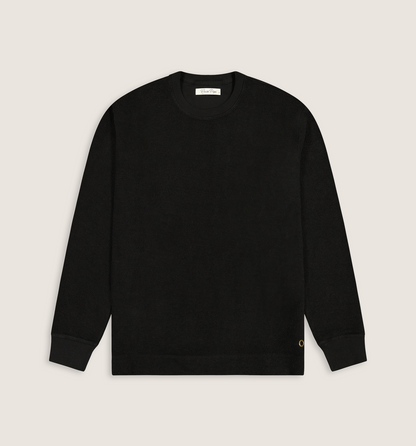 A black long-sleeve crewneck sweater laid flat against a light beige background. A small gold embroidered 'C' logo is visible on the lower left hem, and a white brand label is printed inside the collar.