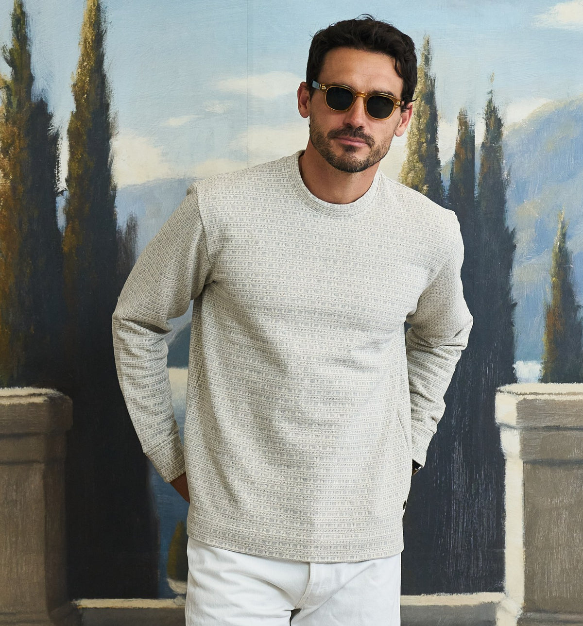 A man wearing a light beige textured knit crewneck sweater and white pants, standing in front of a painted background of cypress trees and mountains.
