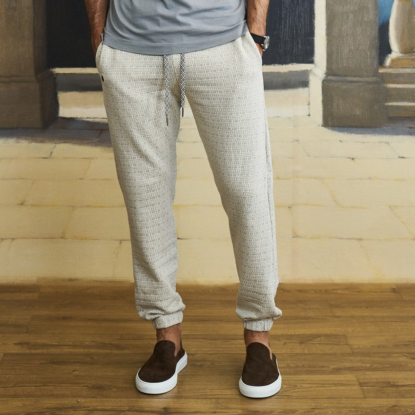 A person wearing light beige textured jogger pants with a drawstring waist, paired with a gray t-shirt and brown slip-on shoes, standing on a wooden floor.